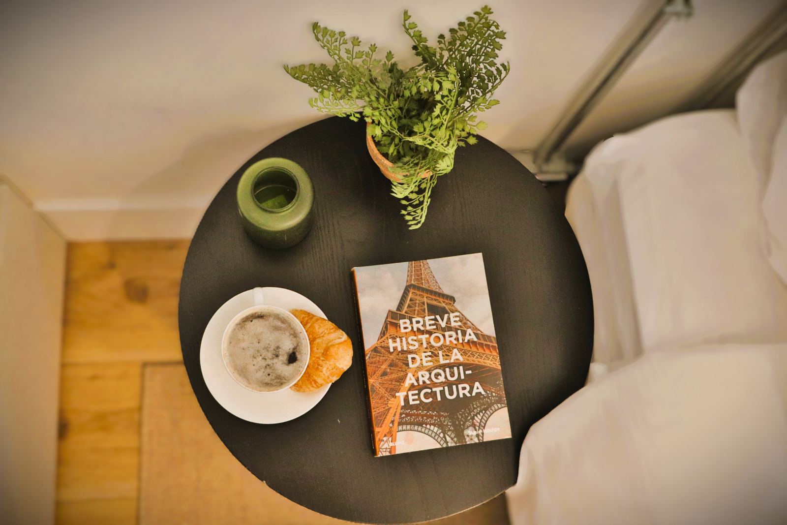 Vista cenital del desayuno servido junto a la cama: café, croissant y libro de arquitectura